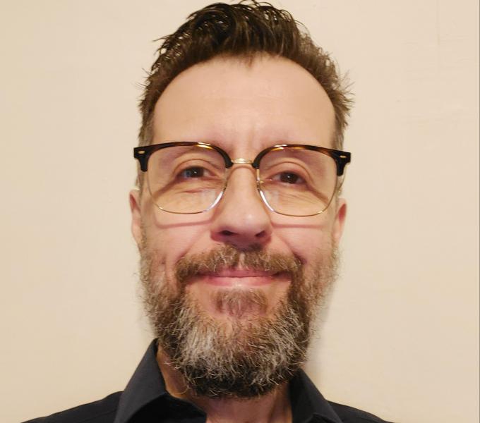 Image of Robert Peckyno.  Image shows a smiling man with a salt & pepper bearded man.  He is wearing a black shirt and brown tortise shell glasses.